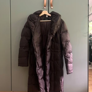 Lång mörkbrun pufferjacka från Esprit - Säljer en lång, mörkbrun pufferjacka från Esprit med huva och dragkedja. Jackan är quiltad och har en clean look som passar perfekt när det är kallt ute. Fodrad och riktigt mysig, med långa ärmar och diskreta detaljer.