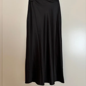 Svart långkjol i satin från NA-KD - Supersnygg svart långkjol från NA-KD i glansigt satinliknande material. Kjolen har en stilren och flowy passform som ger en elegant vibe. Perfekt att styla med chunky sneakers eller boots för en cool look. Aldrig använd!