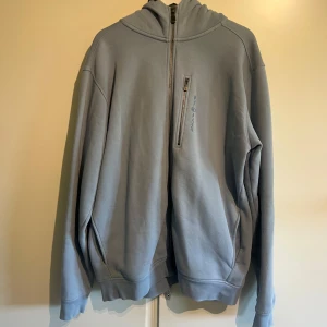Blå hoodie från Sail Racing - Ljusblå hoodie från Sail Racing i storlek 3XL. Tröjan har huva, half zip-dragkedja framtill och en bröstficka med dragkedja. Tillverkad i mjuk bomullsmix och har en relaxed, loose passform. Perfekt för chill dagar eller när du vill ha en clean look.