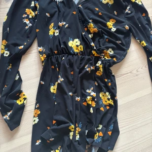 Svart blommig omlottklänning - Supersöt svart kort byxdress med gula och vita blommor, omlottmodell och långa ärmar. Byxdressen har resår i midjan och är perfekt för dig som gillar en feminin och trendig stil. Materialet känns lätt och flowy.