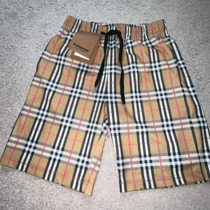 burberry shorts rutiga med snörning  - Snygga Burberry shorts i klassiskt beige rutigt mönster med svarta, vita och röda detaljer. De har elastisk midja med svart snörning för skön passform. Perfekta för sommaren och riktigt stilrena till streetwear. Materialet är mjukt och lätt, troligen bomull eller polyester.