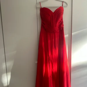 Axelbandlös röd långklänning med slits - Snygg axelbandlös röd långklänning med och draperad överdel. Klänningen har en markerad midja, v-ringad byst och en elegant slits framtill. Tyget är lätt och faller fint, perfekt för bal eller fest. Stilren och iögonfallande design