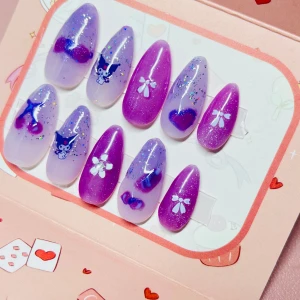 Lila och glitter press-on naglar - Söta press-on naglar i lila och lavendel med glitter, hjärtan, rosetter, blommor och Kuromi-motiv. Naglarna är långa och har en oval form, perfekta för att ge en kawaii vibe till din look. Kommer i set om tio stycken med olika dekorationer.