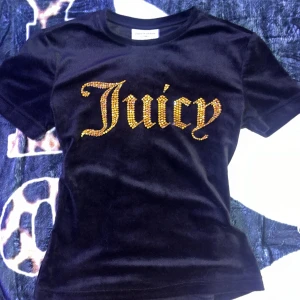 Svart sammetst-shirt Juicy Couture - Svart t-shirt i mjuk sammet från Juicy Couture med korta ärmar. Framsidan har stor guldfärgad text 'Juicy' i glittriga stenar. Perfekt för dig som vill sticka ut med en lyxig och trendig vibe.