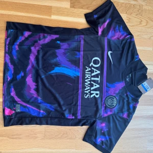 PSG x Nike svart/lila matchtröja - Säljer en fet PSG fotbollströja från Nike med Qatar Airways-tryck. Tröjan är svart med lila och blåa penseldrag över hela framsidan och ärmarna. Klassisk rund hals, kortärmad och tillverkad i lätt, ventilerande material. Perfekt för dig som vill sticka ut på planen eller läktaren.