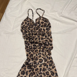 Leopardmönstrad långklänning Big Fashion - Snygg långklänning från Big Fashion med leopardmönster i beige och svart. Klänningen har smala axelband och figurnära passform, perfekt för dig som vill sticka ut. Materialet är glansigt och känns lyxigt mot huden. Supersnygg till klackar och smycken.