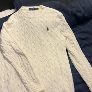 Vit kabelstickad tröja Polo Ralph Lauren - Vit kabelstickad tröja från Polo Ralph Lauren med klassisk rund halsringning och broderad marinblå logga på bröstet. Tröjan är långärmad och har ribbade muddar. Perfekt för en clean och stilren look. Vid frågor eller pris, är det bara att fråga 😄