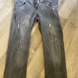 Grå slitna jeans från Catch - Säljer ett par grå jeans från Catch med snygga slitningar och distressed detaljer på benen. Jeansen har klassisk femficksdesign, dragkedja och knapp i midjan. Perfekta för dig som gillar en avslappnad och trendig look.