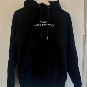 Svart hoodie Peak Performance S - Svart hoodie från Peak Performance i storlek S. Klassisk modell med huva, snörning och magficka. Vit broderad logga på bröstet ger en clean look. Perfekt för chill dagar eller när du vill ha en enkel streetstyle.