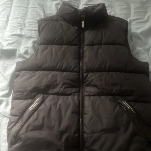 Svart dunväst från Woxo 720 - Svart dunväst från Woxo 720 med hög krage och dragkedja framtill. Västen har två snedställda fickor med dragkedja och quiltad design. Perfekt för lager-på-lager och chill höst- eller vinterstil.