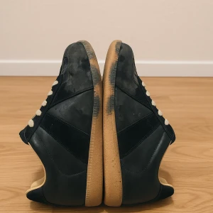 Maison margiela - Snygga svarta sneakers med beige snörning och klassisk gummisula i ljusbrun färg. Ovandelen är i en mix av skinn och mocka, vilket ger en stilren och sportig look. Perfekta för dig som gillar retrovibbar och streetstyle. Helt nya med kvitto