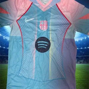 FC Barcelona x Nike kortärmad fotbollströja - Snygg FC Barcelona fotbollströja från Nike med Spotify-logga på bröstet. Tröjan har ett unikt geometriskt mönster med tunna linjer i gult och vitt, och färger i ljusblått, rosa och lila. Röd kant längs sidorna och V-ringad hals. Perfekt för dig som vill sticka ut på planen.