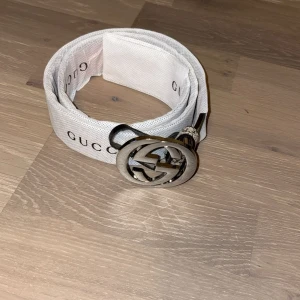 Gucci GG Supreme beige bälte - Snyggt Gucci-bälte i beige med klassiskt GG Supreme-mönster och stor silverfärgad GG-logga som spänne. Bältet är brett och har detaljer i skinn på insidan. Perfekt för att lyfta vilken outfit som helst med en lyxig touch.