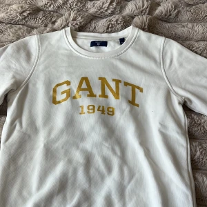 Vit GANT sweatshirt med guldigt tryck XS - Vit sweatshirt från GANT i storlek XS. Tröjan har rund halsringning och långa armar. Tillverkad i mjuk bomull.