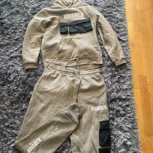 Beige hoodie från Hoodrich med svarta fickor - Snygg beige hoodie från Hoodrich med vita logotyper och två svarta fickor framtill. Tröjan har huva med dragsko och är tillverkad i mjuk bomullsmix. Perfekt för chill dagar och streetwear-vibe.