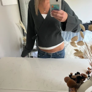 Mörkgrön  omlott kofta med knyt - Snygg mörkgrön omlott kofta med knyt i sidan och breda ribbade muddar. Modellen är croppad och har långa ärmar. 