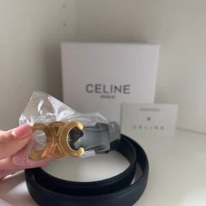 Svart Celine bälte med guldspänne - Stilrent svart bälte från Celine med ikoniskt guldfärgat Triomphe-spänne. Bältet är gjort i slätt skinn och har en klassisk, minimalistisk design som lyfter vilken outfit som helst. Perfekt accessoar för dig som gillar lyxiga detaljer och tidlös stil. Oönskad present så därför inget kvitto kvar