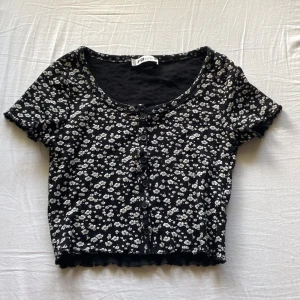 Svart blommig topp från FB Sister - Kortärmad svart topp från FB Sister i storlek XS med vita småblommor över hela tyget. Toppen är croppad, har rund halsringning och knappar framtill. Materialet är mjukt och stretchigt, perfekt för en chill och söt look.