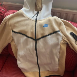 Nike beige och vit hoodiejacka XS - Nike hoodiejacka i beige och vitt med svarta detaljer och blå Nike-logga på bröstet. Jackan har hel dragkedja, huva och en cool ficka med dragkedja på ärmen. Tillverkad i mjuk bomullsmix, perfekt för chill eller streetwear.