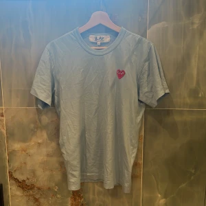 Blå t-shirt Comme des Garçons Play - Snygg ljusblå t-shirt från Comme des Garçons Play i bomull. Klassisk passform med korta ärmar och rund hals. På bröstet finns det ikoniska röda hjärtat med ögon. Perfekt för dig som gillar stilrena plagg med en twist.