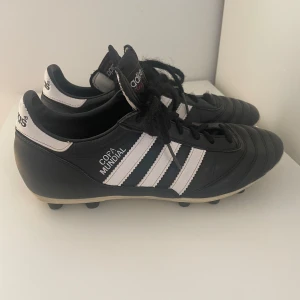 Adidas Copa Mundial fotbollsskor svart/vit - Klassiska Adidas Copa Mundial fotbollsskor i svart skinn med vita detaljer och de ikoniska tre ränderna på sidorna. Skorna har snörning och rund tå, samt dobbar för gräsplan. Perfekta för dig som vill ha stilrena och tidlösa fotbollsskor. Nypris ca 1400