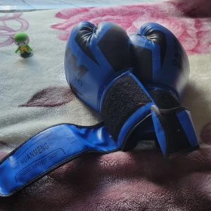 Blå boxningshandskar från HUANMIENG - Säljer ett par blå boxningshandskar från HUANMIENG med svarta detaljer och bred kardborrestängning runt handleden. Handskarna är tillverkade i syntetmaterial och har en sportig look som passar perfekt för träning och boxning.