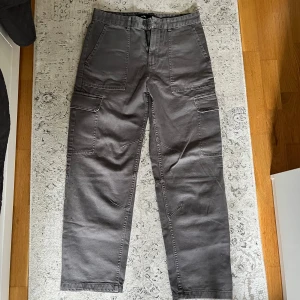 Bershka Cargobyxor - Snygga grå cargobyxor från Bershka med raka ben och stora fickor på sidorna. Byxorna har en avslappnad passform och är tillverkade i bomull, perfekta för en chill och trendig look. Köpta för 280 ZL i Bershka Butik i Polen, sparsamt använda, 280 ZL motsvarar 🟰 690 kronor. Storlek EU38 vilket är typ XS/S