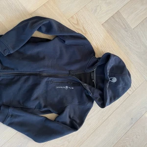 Svart hoodie från Sail Racing - Svart hoodie från Sail Racing med dragkedja och huva. Tröjan har en bröstficka med dragkedja och Sail Racing-logga på bröstet och huvan. Perfekt för dig som gillar sportig och clean stil.