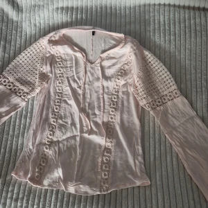 Ljusrosa blus med spets från Freequent - Supersöt ljusrosa blus från Freequent med långa ärmar och v-ringning. Blusen har fina spetsdetaljer längs ärmar och framtill, samt dekorativa band vid halsen. Perfekt för dig som gillar romantisk och feminin stil.