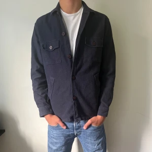 Mörkblå overshirt från John Henric - Snygg mörkblå overshirt från John Henric med strukturerad bomullskänsla. Jackan har två bröstfickor med knapp, sidofickor och knappar framtill. Perfekt lager-på-lager-plagg med klassisk krage och lång ärm. Passar dig som gillar stilren och enkel design. Skriv ifall ni har några funderingar.