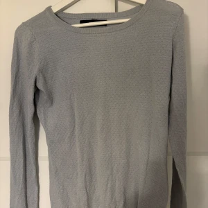 Ljusgrå stickad tröja med struktur - Snygg ljusgrå stickad tröja med diskret strukturmönster. Tröjan har rund hals och långa ärmar, perfekt för dig som gillar enkel och stilren design. Passar till jeans eller kjol och är supermjuk mot huden.