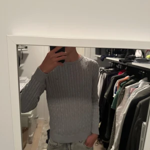 Grå kabelstickad tröja gant - Säljer en grå kabelstickad tröja från gant. Tröjan har rund halsringning, ribbade muddar och diskret logga på bröstet. Perfekt för dig som gillar en clean och klassisk stil. Passar lika bra till jeans som till chinos. Passar dig mellan 165-175cm
