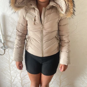 Beige pufferjacka med fuskpäls från Hollies - Snygg beige pufferjacka från Hollies med stor huva och fluffig fuskpäls i bruna och svarta toner. Jackan har dragkedjor i guld, flera fickor och en badge på ärmen. Perfekt för kalla dagar och ger en riktigt trendig look. Storlek 34 som motsvarar S/M, och knappt använd. ABS! Pris går att diskutera.