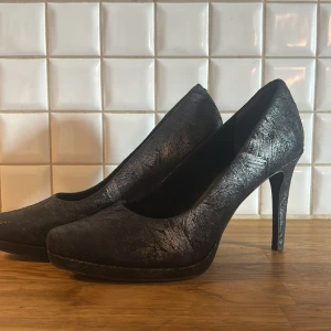 Svarta högklackade Tamaris skor - Snygga svarta högklackade skor från Tamaris med rund tå. Skorna har en cool textur i materialet och fyra silvriga nitar bak på hälen som ger extra edge. Perfekta för dig som vill sticka ut lite extra. Klassiskt snitt och plattform framtill för bekvämare känsla.