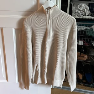 Beige stickad half zip från J.Lindeberg - Snygg beige stickad tröja från J.Lindeberg med ribbad struktur och half zip-dragkedja i halsen. Perfekt för dig som gillar stilrena och moderna plagg. Tröjan har lång ärm och hög krage för extra komfort.
