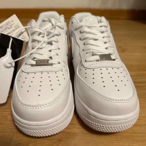 Nike Air Force 1 helvita sneakers - Klassiska Nike Air Force 1 i helvitt skinn med perforerad tå, vit sula och ikonisk Swoosh på sidan. Lågt skaft, snörning och metallbricka med 'AF1' på snörena. Perfekta för en clean och stilren look.