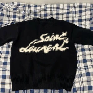 Black designer hoodie M - Svart stickad tröja från Saint Laurent i storlek M. Tröjan har en oversized look med bred ribbad mudd och ett stort, fluffigt vitt Saint Laurent-tryck framtill. Perfekt för dig som gillar statement-plagg och vill sticka ut.