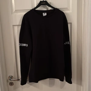 Svart sweatshirt med tryck URB4N - Svart sweatshirt från Urban Classics med stora vita tryck på ryggen och textdetaljer på ärmarna. Tröjan har rund hals, lång ärm och en loose passform. Perfekt för dig som gillar streetstyle och vill sticka ut med grafiska detaljer.