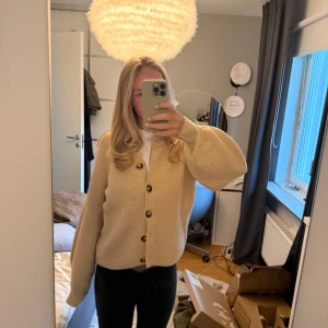 Beige stickad kofta med knappar - Säljer en beige stickad kofta med stora bruna knappar framtill. Koftan har en loose passform och långa ärmar med lätt ballongeffekt. Perfekt att slänga på över en t-shirt för en chill och snygg look. Supermjuk och skön att bära.