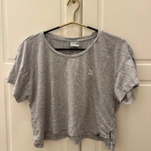 Grå croppad t-shirt från Puma - Grå croppad t-shirt från Puma med kort ärm och rund halsringning. T-shirten har en liten vit Puma-logga på bröstet och en diskret etikett nertill, perfekt för en chill och sportig look.