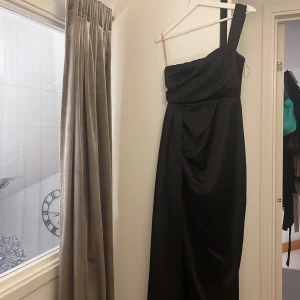 Svart långklänning med oneshoulder - Snygg svart långklänning med oneshoulder och draperad detalj framtill. Klänningen har en elegant slits och är gjord i ett glansigt, mjukt material som ger ett lyxigt intryck. Perfekt för dig som vill sticka ut med en stilren och modern look.