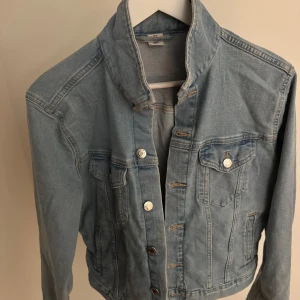 Ljusblå jeansjacka med knappar - Snygg ljusblå jeansjacka med klassisk krage och silverfärgade knappar framtill. Jackan har två bröstfickor med lock och knapp samt två sidofickor. Perfekt för lager-på-lager och enkel att styla till olika outfits.