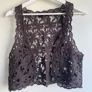 Virkad väst med blommönster - Säljer en grå/brun virkad väst med blommiga detaljer och öppen struktur. Västen har en kort, croppad passform och är perfekt för att ge en bohemisk touch till din outfit. 