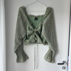 Grön blommig blus från Vero Moda - Supersöt blommig blus från Vero Moda i ljusgrönt med små röda och rosa blommor. Blusen har långa puffiga ärmar med smock och volang vid ärmslut, v-ringning, smockad kropp och snörning framtill. Perfekt för vår och sommar när du vill ha en romantisk look.