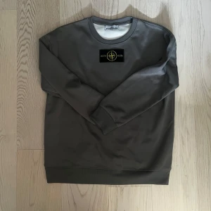 Grå Stone Island sweatshirt - Snygg grå sweatshirt från Stone Island med klassisk rund hals och ribbade muddar. Tröjan har den ikoniska Stone Island-patchen i svart, gul och grön på ärmen. Perfekt för dig som gillar streetwear och vill ha en clean look med tydlig logga.