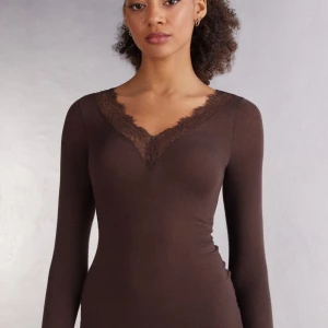 Brun långärmad topp med spets - Mörkbrun långärmad topp med v-ringning och fina spetsdetaljer längs halsringningen och vid ärmsluten. Toppens material är mjukt och stretchigt, vilket ger en slim passform. Perfekt för dig som gillar en stilren look med en touch av romantik.