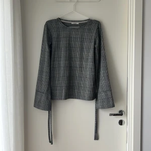 Rutig långärmad tröja från Pieces - Snygg svartvit rutig tröja från Pieces i storlek M. Tröjan har långa ärmar med coola knytband vid ärmsluten som ger en unik touch. Rak passform och rund halsringning. Perfekt för dig som gillar stilrena och trendiga plagg.