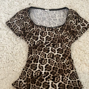 Leopardmönstrad topp från Stories XS - Supersnygg leopardmönstrad topp från Stories i storlek XS. Topp med kort ärm, figurnära passform och söt svart spetskant vid den runda urringningen. Tunt och stretchigt material som känns lätt på kroppen. Perfekt för dig som vill sticka ut med ett djurmönster.