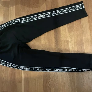 Svarta mjukisbyxor Emporio Armani L - Svarta mjukisbyxor från Emporio Armani med vita loggor längs sidorna och broderad logga framtill. Byxorna har elastisk midja, dragkedjeförsedda fickor och är tillverkade i mjukt material. Perfekta för en chill och sportig look.