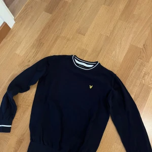 Mörkblå tröja från Lyle & Scott - Mörkblå tröja från Lyle & Scott med klassisk gul logga på bröstet. Rund halsringning med vita ränder och matchande detaljer vid ärmsluten. Långärmad och i mjukt bomullsmaterial, perfekt för en stilren och sportig look.
